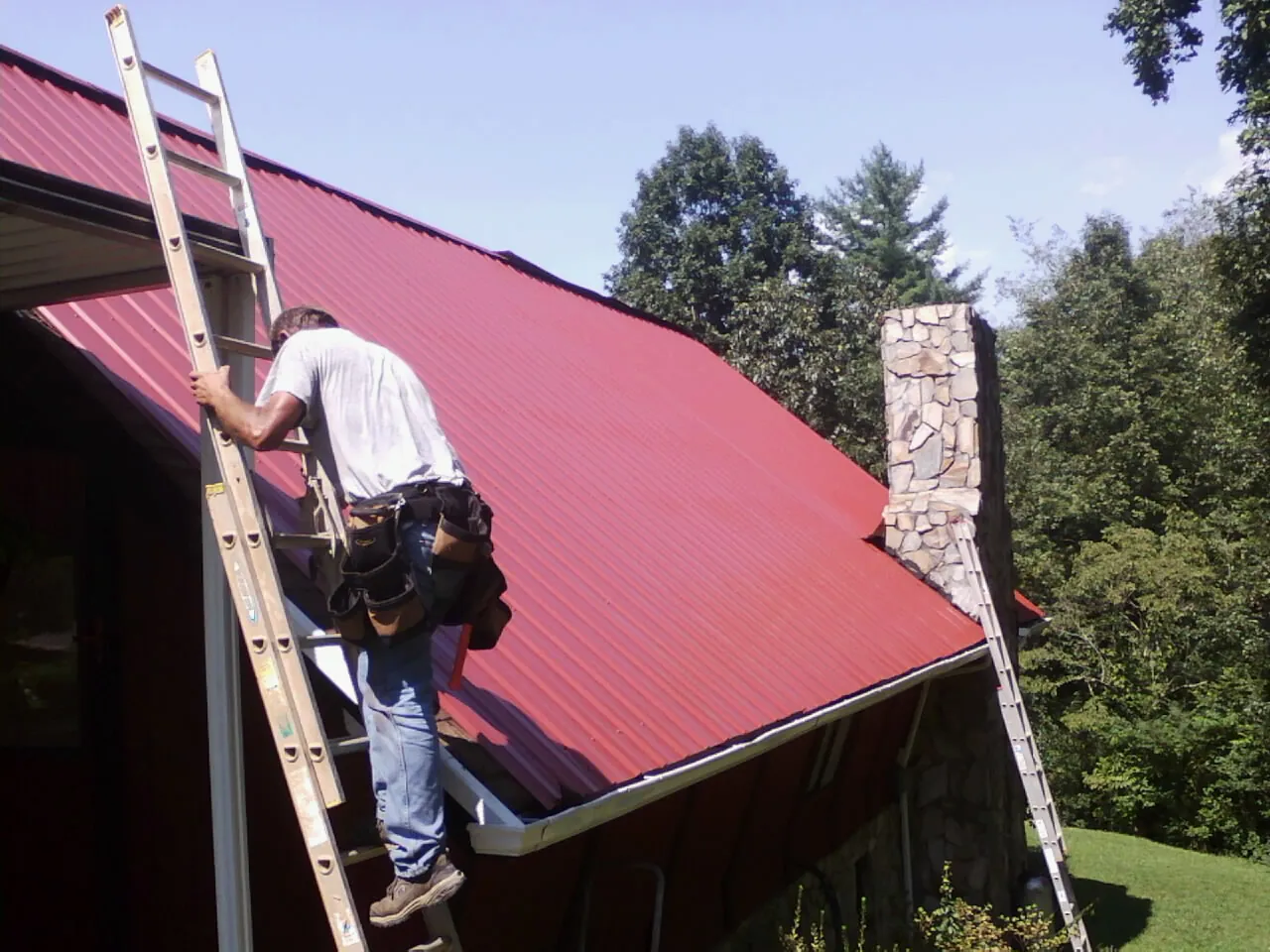 Roofer on a ladder beside a red metal roof and stone chimney — Soffit & Fascia Repair by Roofing Brothers in San Marino